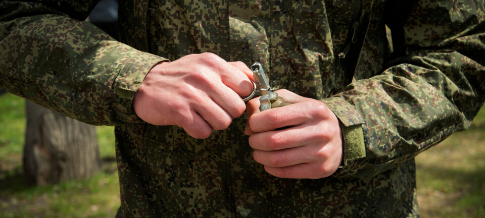 Camouflaged soldier holding a hand grenade with both hands outdoors.