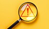 Magnifying glass over a red warning triangle with an exclamation mark on a yellow surface.